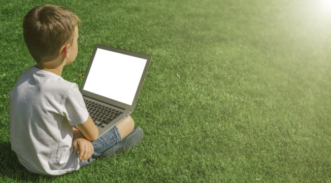 A child sits on the grass and studies with a laptop online, enjoying nature - Powered by Adobe