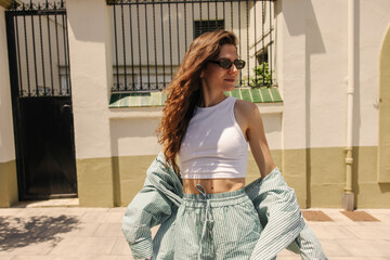 Pretty young caucasian woman in sunglasses looks away, spends time outdoor in sunny weather. Brunette wears white singlet and striped green suit. Concept solo vacation.