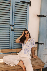 Fototapeta premium Beautiful young caucasian woman sitting on wooden bench and looking at camera on street. Brunette wears sunglass and casual clothes. Relaxation concept.