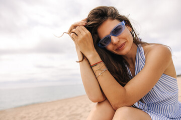 Stylish young caucasian girl posing and looking at camera outside. Brown hair model sits on beach in cloudy weather. Concept tourist, summer holidays.