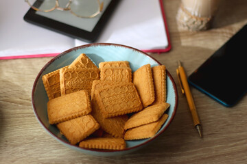 Notebook, pen, tablet, reading glasses, books, phone, candle, bowl of biscuits and cup of tea on the table. Selective focus.