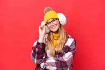 Young beautiful woman wearing winter muffs isolated on red background with glasses and happy