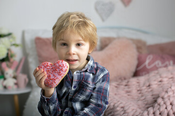 Cute little blond child, preschool boy, eating pink donut in the shape of heart, made for Valentine