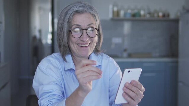 Active mature lady in stylish eyeglasses looking performance of favourite music band in internet on smartphone and dancing, having fun and enjoying, good mood