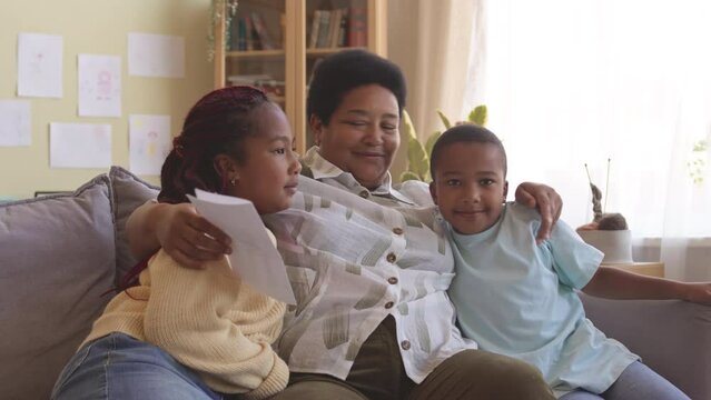 Two nice African American tween kids giving handmade postcard to their grandmother, embracing and kissing them on cheeks