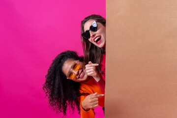 Two smiling beautiful girl friends hold a cardboard sign on a pink background
