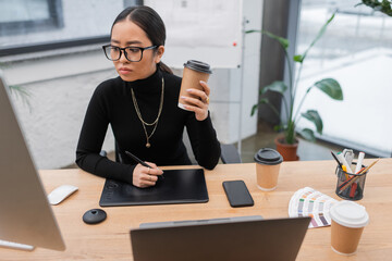 Young asian designer holding coffee to go near graphic tablet and gadgets in office.