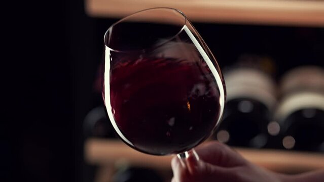 Close up female hand swirling red wine in wine glass. Wine expert tasting, rating and drinking wine, bottles in background. Slow motion video.