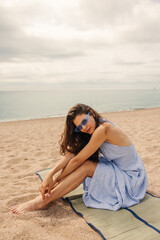 Modern young caucasian woman in blue sunglasses sits on underlay with sea background. Brunette hair model spends time on beach and looking at camera. Concept summer vacation, lifestyle.