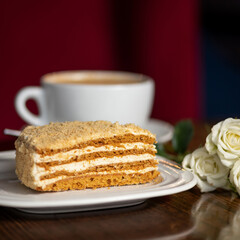 Piece of honey cake on plate with cup of coffee on wooden table. Cutaway honey cake. Table is decorated with roses. Serving dish. Dark red background. 