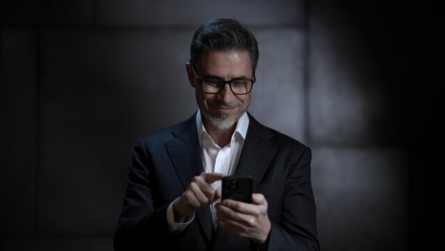 Portrait of happy mature businessman, with phone. Entrepreneur standing in front of dark loft wall. Older, middle aged, mid adult, man in his 40s or 50s in shirt and jacket, business casual.