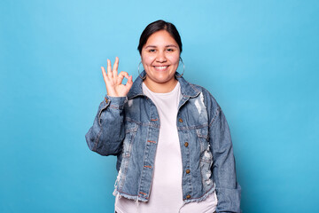 Young curvy latina woman wearing denim jacket and hoop earrings, smiling showing okay sign looking at camera isolated on blue background. Copy space. - obrazy, fototapety, plakaty