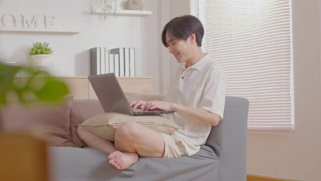 Handsome Asian young man sit on couch looking on laptop screen recieved good news raise hand up celabrating. Happiness Freelancer using computer laptop working online at home. Work from home concept