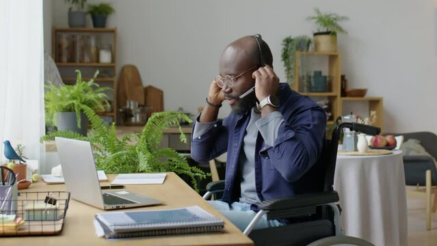 African American man in wheelchair putting on wireless headset and speaking via web call on laptop while working online from home