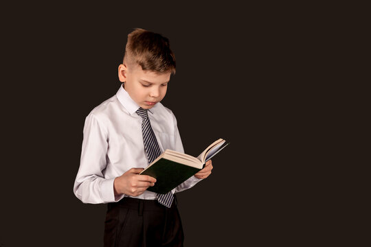 Positive boy standing and reading book with smart expression face, wear white shirt and necktie on black isolated background. Schoolboy posing in studio. Education concept. Copy text space for ad