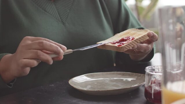 Unrecognizable mature woman using knife while putting delicious strawberry jam on fresh toast during breakfast
