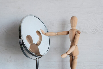 A mannequin wooden man stands on a light background and looks in the mirror. - obrazy, fototapety, plakaty