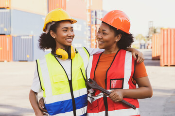 Two african american female workers who match smiling buddy in hard hat works at container terminal...