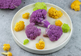 Yellow, purple and green fresh cauliflower inflorescences on white plate on grey background. 
Multicolored food close-up. Raw rainbow cauliflower prepared to be cooked or ready to eat as raw food diet