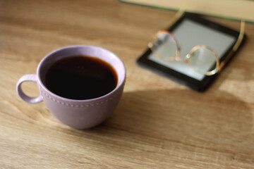 Cup of tea or coffee, e-reader and reading glasses on the table. Pile of books in the background. Selective focus.