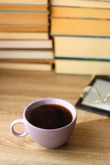 Cup of tea or coffee, e-reader and reading glasses on the table. Pile of books in the background. Selective focus.