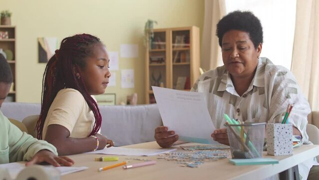Mature Black woman checking home assignment of her granddaughter while sitting together at desk with jigsaw puzzle on it