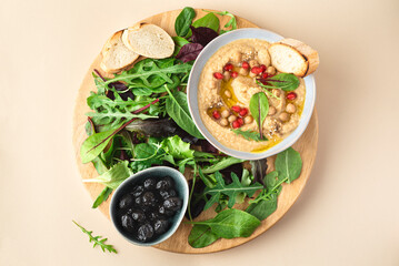 Hummus with pomegranate seeds and various mini greens, arugula, small spinach leaves served with toasted bread and black dried olives. Neutral beige background. Plant based diet. North African cuisine