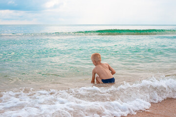 Fototapeta na wymiar Blond boy playing in the sea water on a sandy beach on a summer day, back view. Fun vacation.