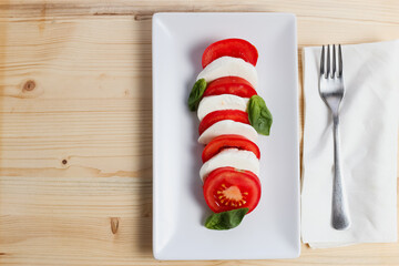 Top view of a fresh caprese salad over a white rectangular plate and a fork over a wooden table