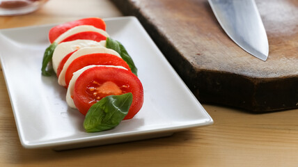 Close up view of a fresh caprese salad over a white rectangular plate, kitchen knife and a bowl of fresh tomatoes over a wooden table