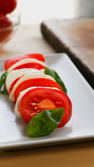 Vertical close up view of a fresh caprese salad over a white rectangular plate, kitchen knife and a bowl of fresh tomatoes over a wooden table