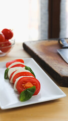 Vertical view of a fresh caprese salad over a white rectangular plate, kitchen knife and a bowl of fresh tomatoes over a wooden table