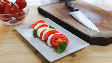 Fresh caprese salad over a white rectangular plate, kitchen knife and a bowl of fresh tomatoes over a wooden table