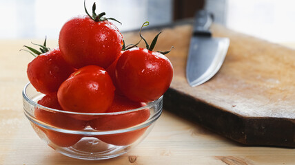Close up of tomatoes on a glass bowl and kitchen knife over a wooden table in front of a window