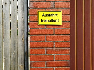 Gelbes Schild aus Plastik Ausfahrt freihalten auf braunem Backstein eines Pfosten neben einem Hoftor in Wettenberg Krofdorf-Gleiberg bei Gießen in Hessen - obrazy, fototapety, plakaty