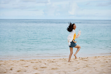 Fototapeta premium Sports woman runs along the beach in summer clothes on the sand in a yellow T-shirt and denim shorts white shirt flying hair ocean view, beach vacation