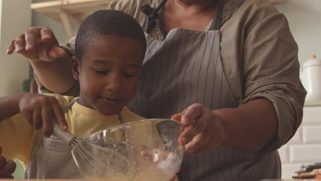 Mature Black woman teaching her little grandson in apron to whisk eggs in glass bowl while making dough at home