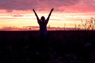  Silhouette of a girl in a field with raised hands against the sunset sky