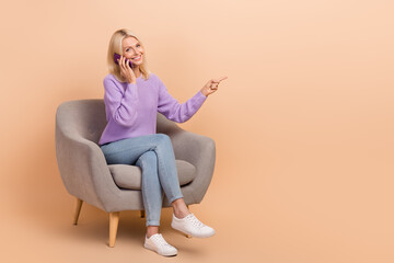 Fototapeta na wymiar Full size photo of nice woman dressed purple sweater jeans on armchair talking directing empty space isolated on beige color background