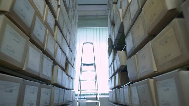 Archive metal racks with boxes lying on shelves and stepladder standing against window at bright sunlight at aisle end motion along passage