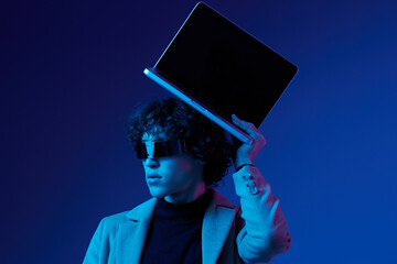 Portrait of a stylish male hacker with a laptop and futuristic glasses in blue light, Blue Perennial color, cyber security, technology and education, neon color, freelancer online work, trend color