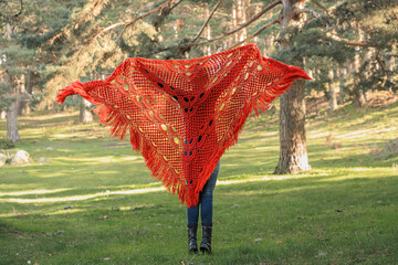 A woman in a red knitted shawl stands in a forest clearing and looks ahead.