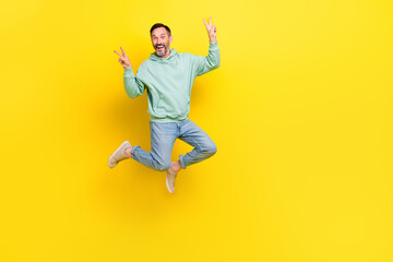 Fototapeta na wymiar Full length photo of funky impressed guy dressed green hoodie jumping high showing v-sign empty space isolated yellow color background