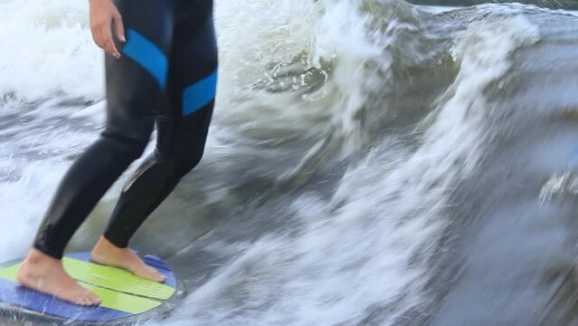 Sportsman dressed in neoprene swimsuit wakesurfs actively at high speed standing on colorful board among waves on river closeup