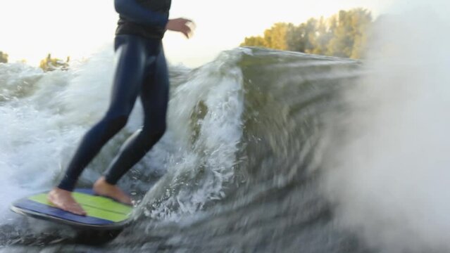 Strong sportsman in neoprene wakesurfs at high speed standing on surfboard among boat trace waves against autumn forestry bank closeup