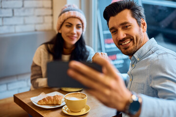 Happy man using mobile phone while taking selfie with his girlfriend in cafe.