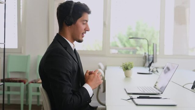Young man wearing wireless headset using a laptop computer for communication with customers or colleagues sitting at the desk in contemporary office. Maale support worker talks online. Side view