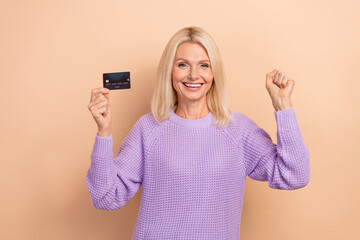 Photo of overjoyed positive retired lady blond hairdo wear knit sweater hold plastic card clench fist isolated on beige color background