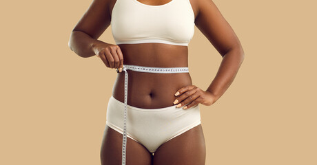 Plus size woman holding measuring tape on waist on beige color background, cropped shot. Beautiful young black female with belly fat and sexy curvy figure planning liposuction for imperfect body areas