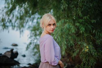 Obraz premium Blonde girl on a summer day on the shore near the river in nature in a dress